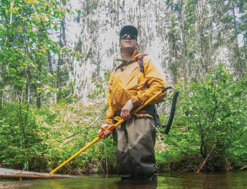 Sam Bourret, biologist with Montana Fish, Wildlife, and Parks, in a yellow jacket standing in a stream.