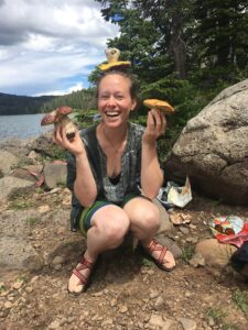A smiling person holds up mushrooms and balances one on her head with a lake in the background. 