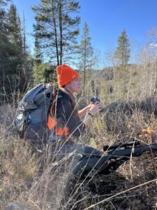 A person in camo and blaze orange with a backpack, binoculars, and a rifle crouches in the grass.