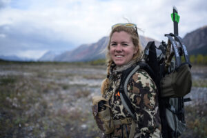 A woman wearing camo and a backpack with a bow and arrows stands before a cloud-draped mountain.