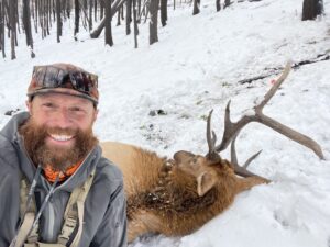 Bearded man smiles at camera in front of a downed elk in the snow.