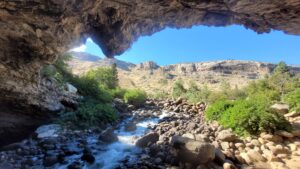 A view from inside a cave looking out to blue skies and cliff walls, with a river framed by green vegetation.
