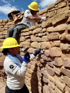 Two people in hard hats holding hammers work on a stacked stone wall. 