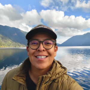 Photo of man wearing glasses and a hat in front of a mountain lake.