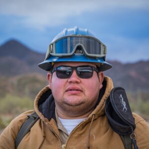 Photo of man wearing sunglasses and a hardhat with mountains behind.
