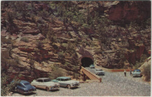 An old postcard of Arches National Park that shows a number of vintage cars parked outside a tunnel through canyon walls.