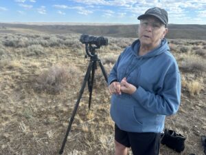 A woman stands in sagebrush next to a tripod-mounted camera.